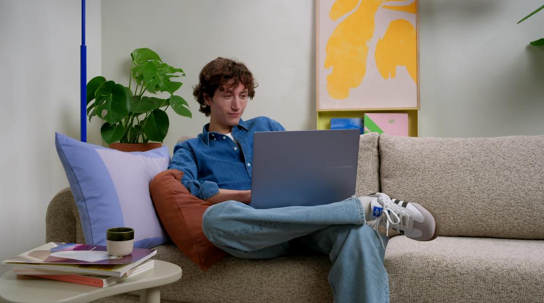Jovem sentado em um sofá, usando um laptop prateado em um ambiente iluminado e decorado.