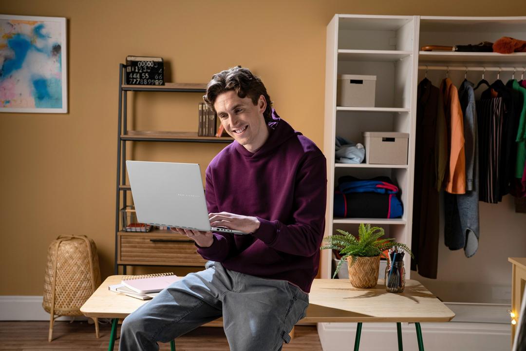 Homem sorridente sentado em uma mesa, utilizando o ASUS Vivobook S 14, em um ambiente aconchegante com prateleiras e roupas ao fundo.