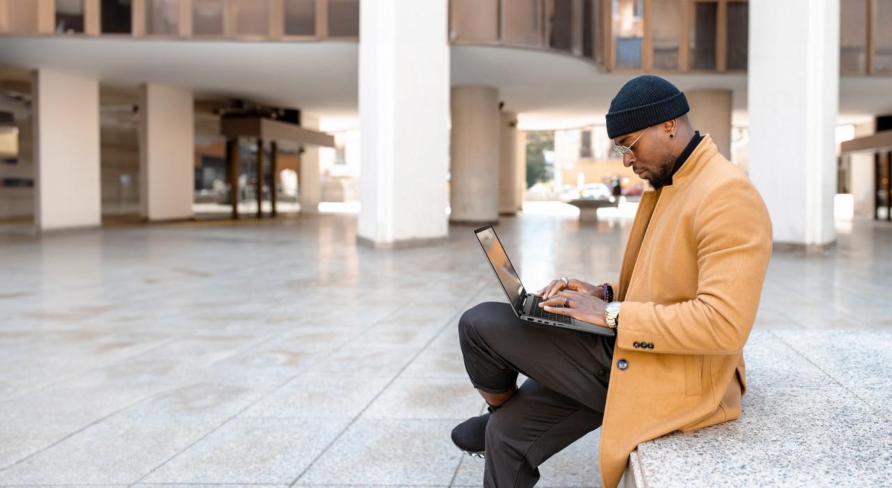 Homem usando um notebook ao ar livre, vestindo casaco bege, ideal como presente para o dia dos pais, simbolizando modernidade e praticidade.