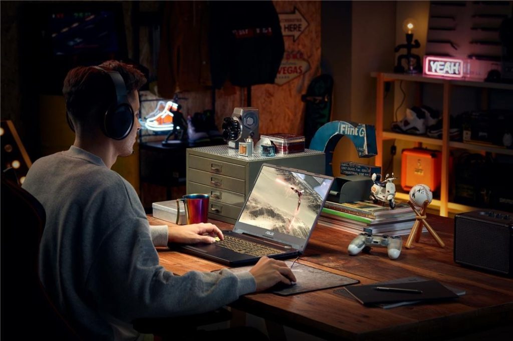 Pessoa jogando em um notebook gamer ASUS TUF Gaming A15 em uma mesa organizada, com fones de ouvido e itens de decoração ao redor.
