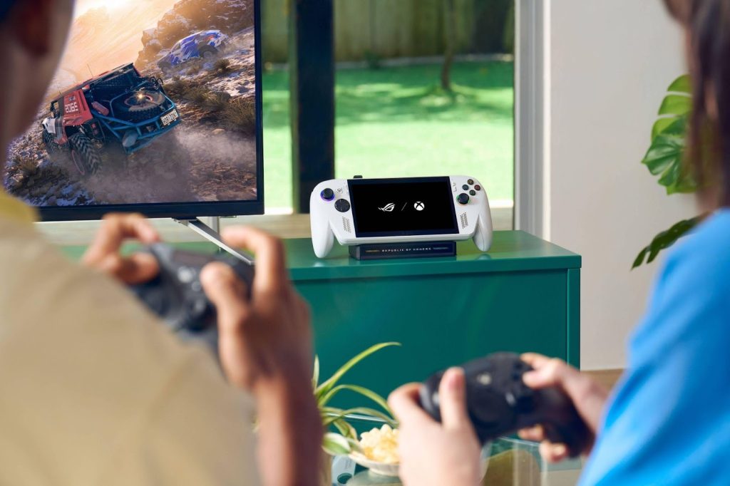 Duas pessoas jogam com controles; ao fundo, um console portátil sobre um móvel verde e uma TV exibindo um jogo de corrida.