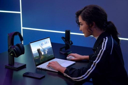Uma jovem de cabelos escuros e jaqueta esportiva utiliza um notebook ASUS ROG Zephyrus branco em uma mesa de setup gamer.