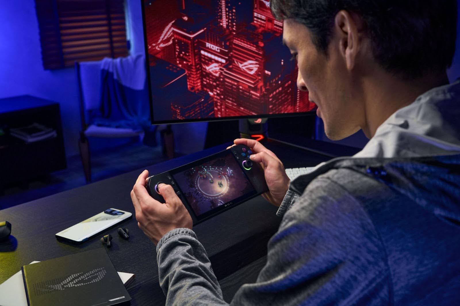 Pessoa jogando em um console portátil ROG Ally X em uma mesa gamer, com monitor e smartphone ASUS ROG ao fundo.