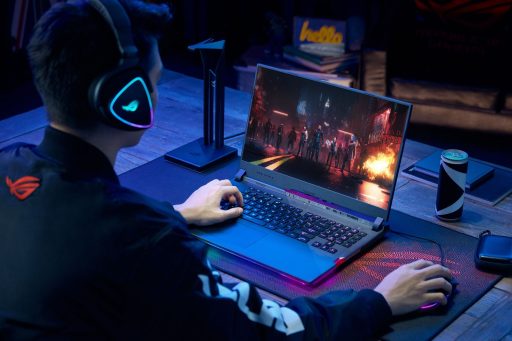 Gamer usa headset e notebook ROG Strix SCAR 17 com iluminação RGB para jogar em ambiente escuro sobre mousepad temático e mesa de madeira.