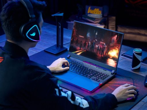 Gamer usa headset e notebook ROG Strix SCAR 17 com iluminação RGB para jogar em ambiente escuro sobre mousepad temático e mesa de madeira.