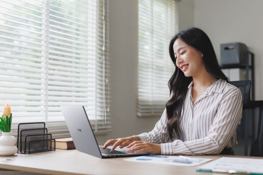 Mulher de camisa listrada sorrindo enquanto digita em um notebook ASUS Expertbook cinza, sentada em um escritório iluminado próximo a uma janela com persianas.