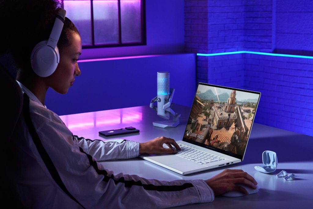 Mulher com headphone branco joga no notebook ROG Zephyrus G16 em ambiente com iluminação neon roxa, mostrando interface do Game Pass na tela.