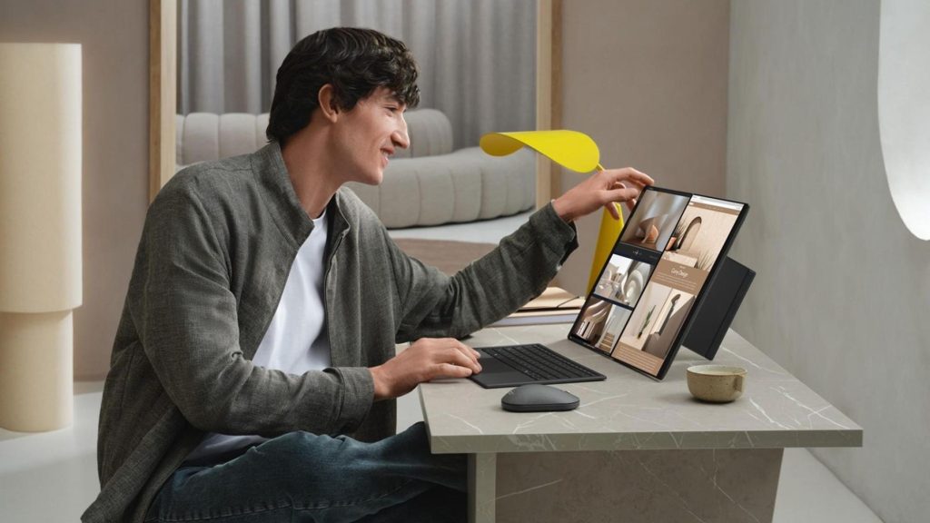 Homem sorrindo ao utilizar um notebook ASUS de tela dupla posicionado verticalmente sobre uma mesa de mármore, com teclado e mouse sem fio.