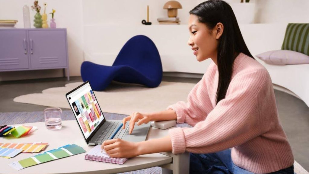 Mulher sorrindo sentada diante de um notebook ASUS que exibe paletas de cores na tela, enquanto faz anotações em um caderno com amostras de design sobre a mesa.