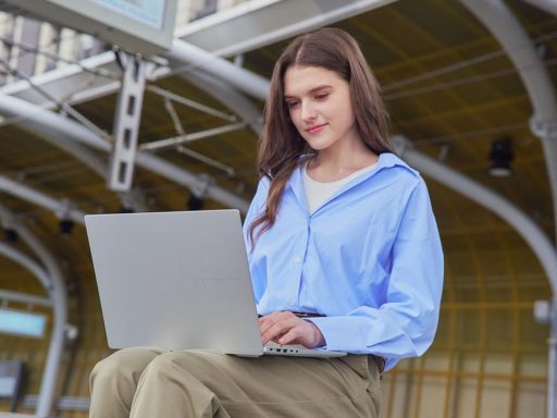 Mulher sentada em ambiente externo moderno utilizando um ASUS notebook prata. Ela veste camisa azul e calça bege, com foco na tela do aparelho.