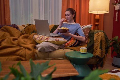 Jovem sentada confortavelmente em um sofá aconchegante come pipoca enquanto assiste a vídeos no notebook ASUS Vivobook S14.