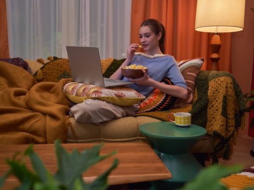 Jovem sentada confortavelmente em um sofá aconchegante come pipoca enquanto assiste a vídeos no notebook ASUS Vivobook S14.