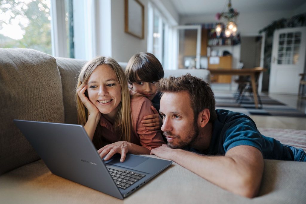 Família deitada no sofá reunida e sorrindo enquanto olham juntos para a tela de um notebook Vivobook 16 de cor cinza escuro na sala de casa.