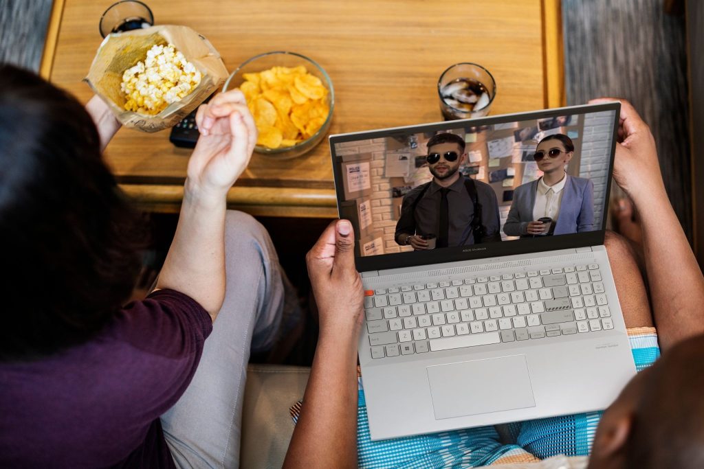 Duas pessoas vistas de cima consomem pipoca e batatas enquanto assistem a um filme de ação na tela de um notebook ASUS Vivobook Pro 15X OLED.